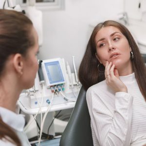 woman in dental chair holding jaw and speaking to dentist